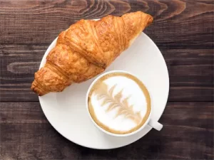 Fotografija rogljička in kave z mlekom na krožniku na leseni podlagi.